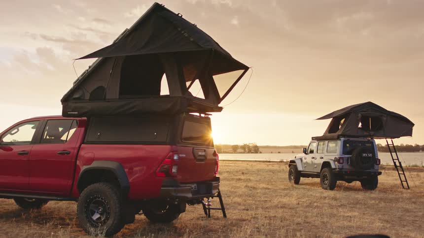 Two rugged off-road vehicles with rooftop tents are set up for an overland adventure in a field by a lake at golden hour