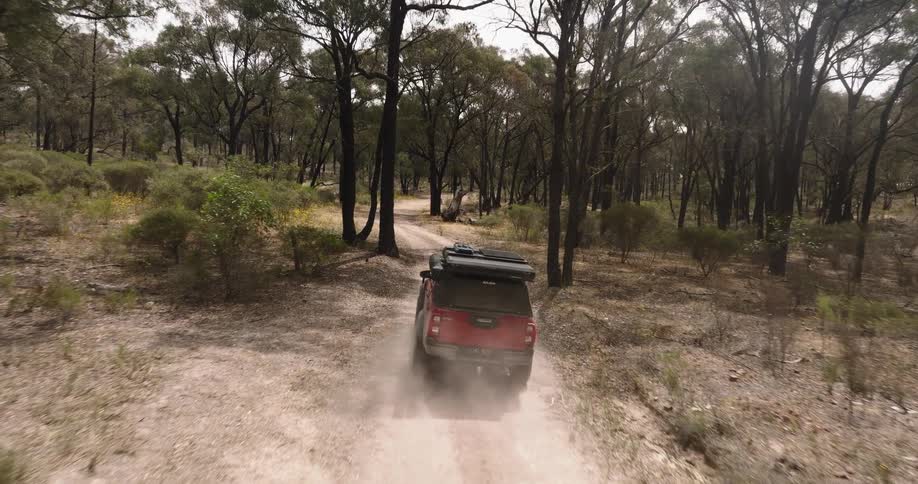 An aerial view follows a red off-road truck with a camper as it drives down a dusty dirt road through a dry forest