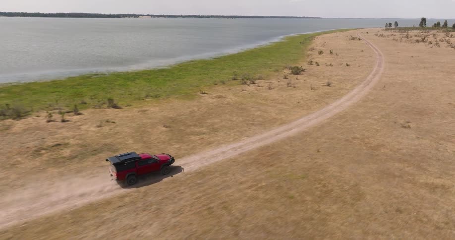 Aerial slow-motion shot of a red pickup truck off-roading on a dusty dirt track alongside a vast lake on a sunny day