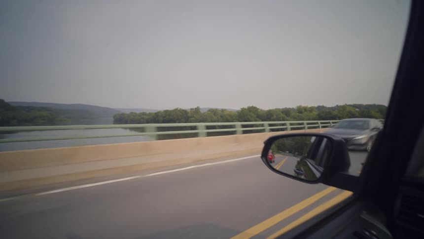A driver's point of view shot from inside a car driving across a long bridge over a wide river on a hazy day