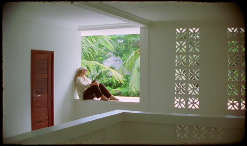 A woman sits peacefully in an open-air window of a white tropical building, looking out at the lush green palm trees