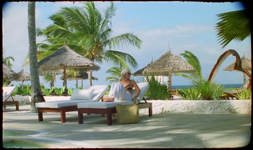 Vintage-style footage shows a woman relaxing on a sun lounger at a luxurious tropical beach resort