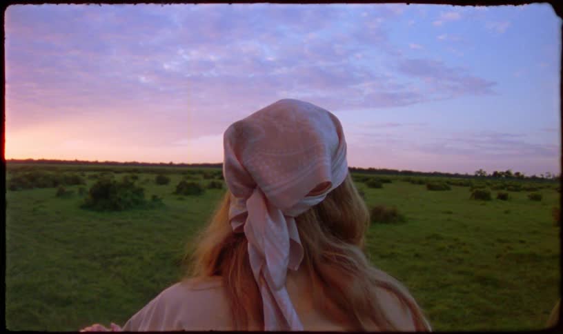 Vintage-style footage shows a woman from behind looking at a beautiful pink and purple sunset over a green field