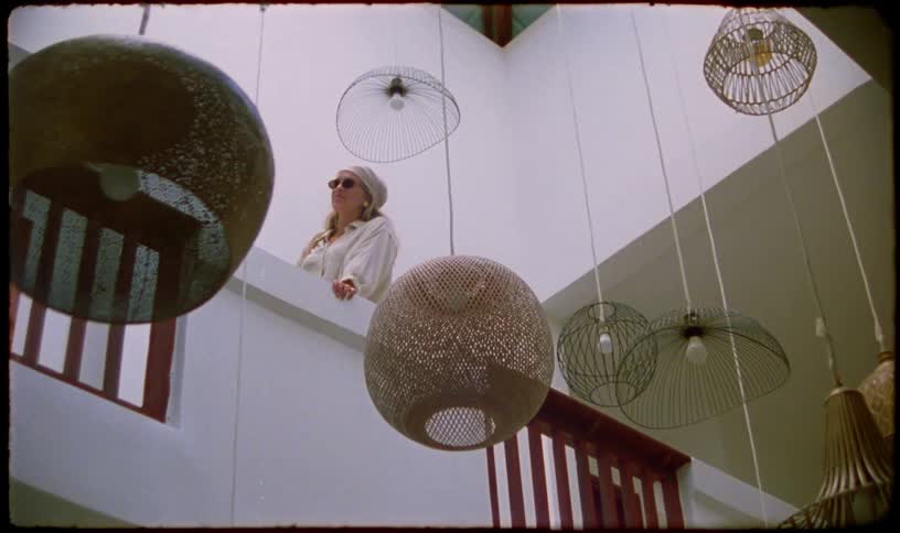 A low-angle shot captures a stylish woman in sunglasses and a headscarf looking out from a modern balcony, with artistic woven lampshades hanging in the foreground
