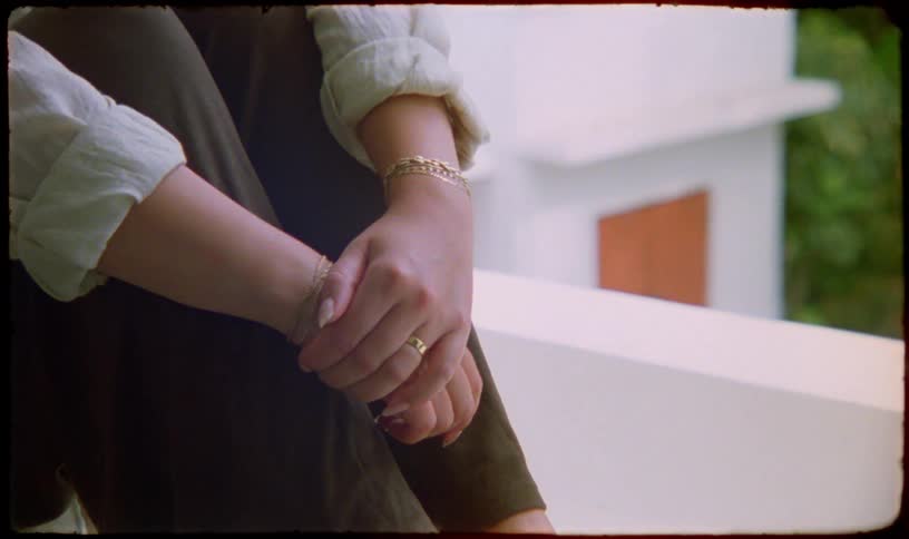 Close-up on the hands of a woman wearing gold jewelry as she sits pensively on a balcony in a vintage-style shot