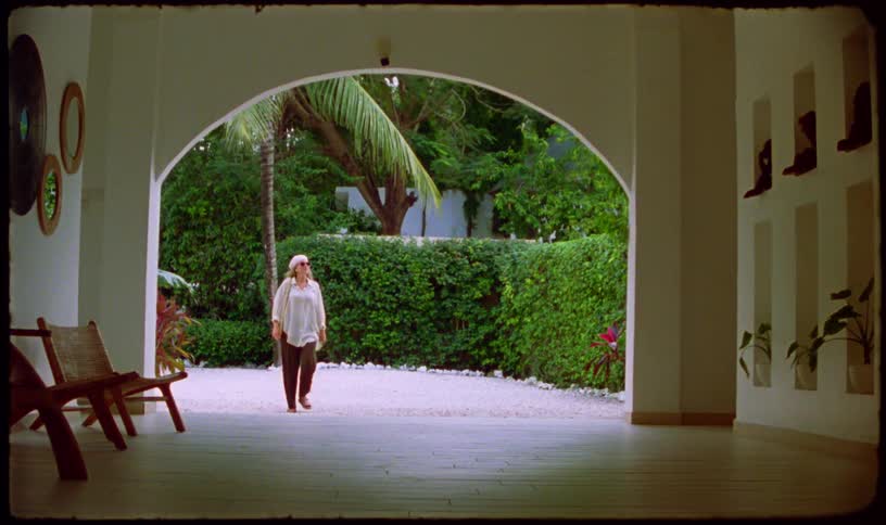 A stylish woman in sunglasses walks from a lush tropical garden into a modern villa, captured on vintage-style film