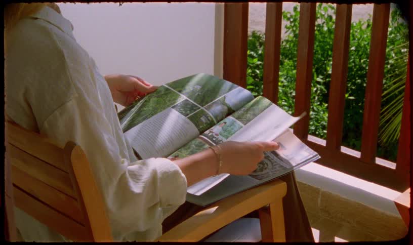 A woman leisurely flips through a magazine while relaxing in a chair on a sunlit balcony, captured in a warm, nostalgic film style with a light leak effect