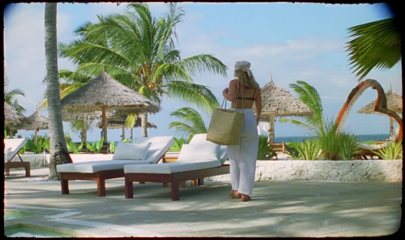 A blonde woman in chic beach attire walks towards lounge chairs at a sunny tropical resort with a vintage film aesthetic