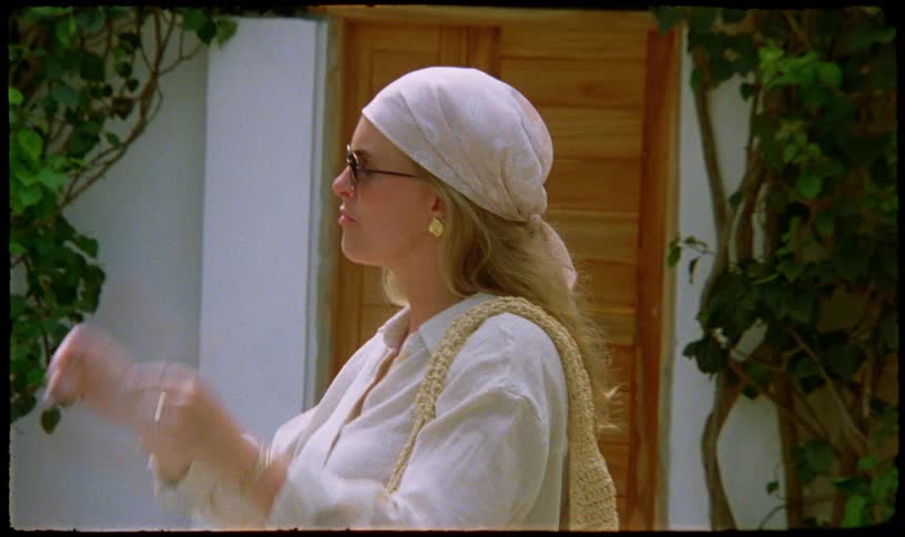 A stylish blonde woman in a headscarf puts on her sunglasses while standing in a sunny, vine-covered courtyard
