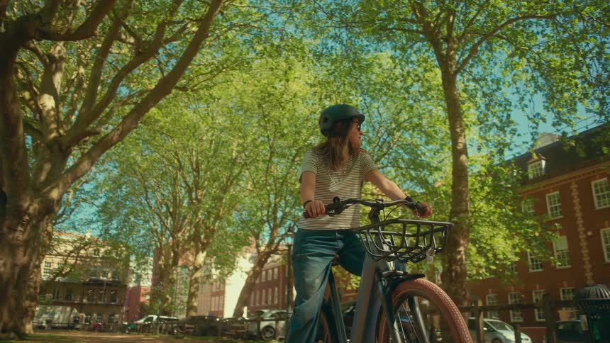 A woman wearing a helmet and sunglasses pauses on her bicycle before riding through a sun-drenched, tree-lined city street