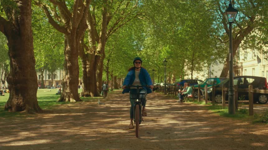 Slow motion shot of a woman riding an e-bike towards the camera on a sunlit path in a London park