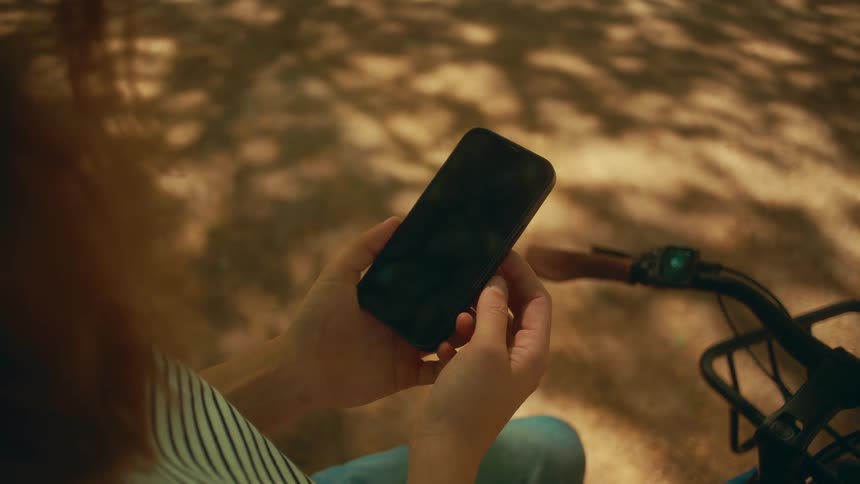 Close-up over-the-shoulder shot of a person holding a smartphone with a blank screen next to their bicycle in a sun-dappled park