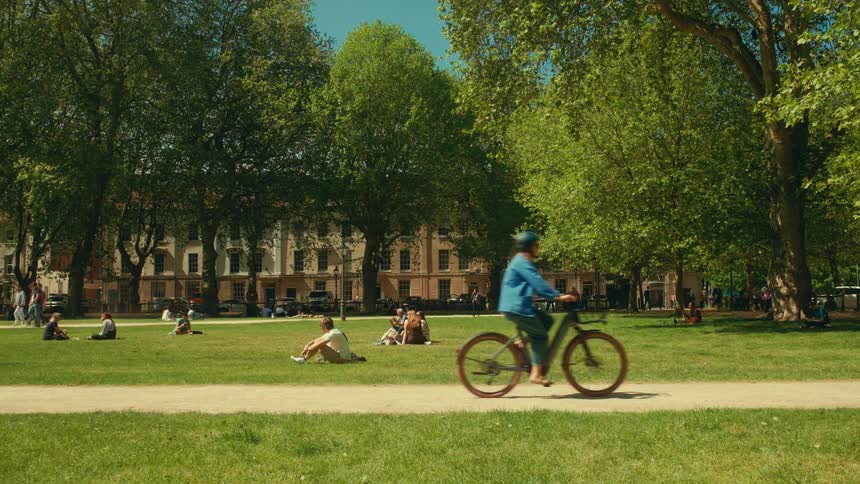 A cyclist rides past people relaxing and enjoying a sunny day in a lush green city park