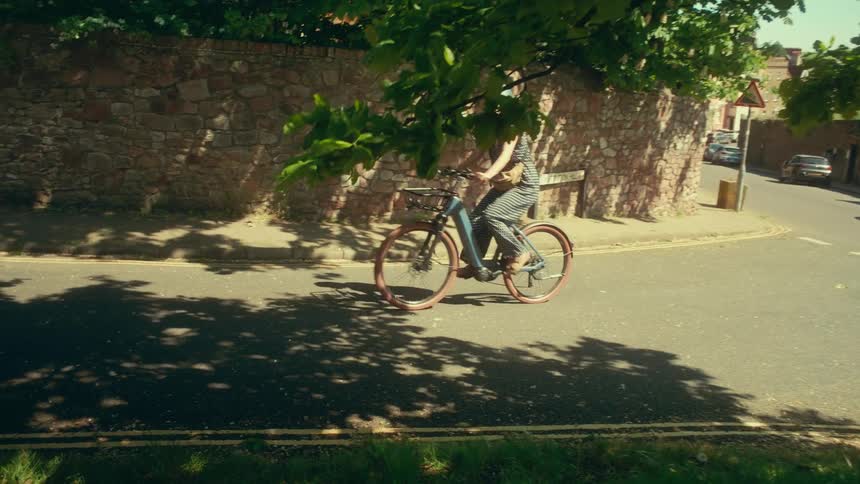 A woman in a helmet rides her electric bike down a quiet, sun-dappled town street