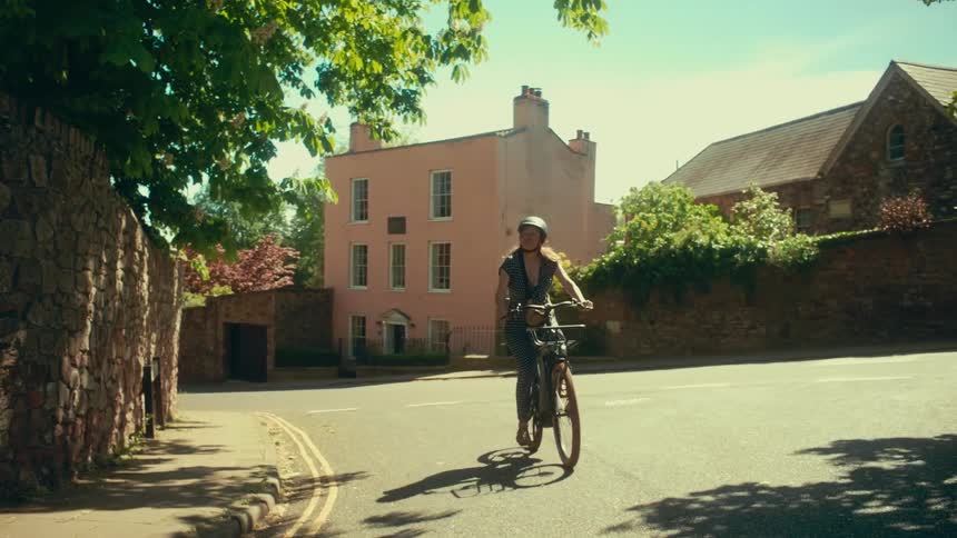 A woman wearing a helmet cycles in slow motion down a sun-drenched street in a quaint town
