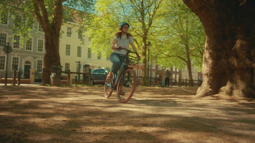 Slow motion shot of a young woman cycling through a tree-lined city square on a beautiful sunny day