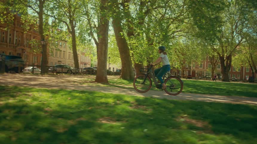 A woman enjoys a leisurely ride on her electric bike through a sun-dappled city park as joggers run nearby