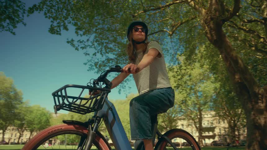 Low angle tracking shot of a woman wearing a helmet and sunglasses riding an e-bike through a park on a sunny day