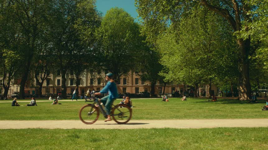 A cyclist rides past people relaxing and sunbathing on the lush green grass of a city park on a beautiful sunny day