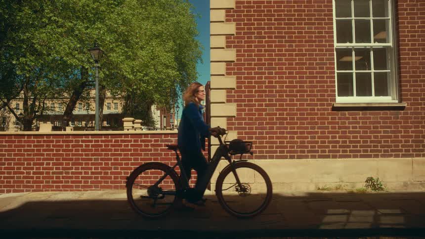 A woman walks her electric bicycle down a city sidewalk past a brick wall on a sunny day