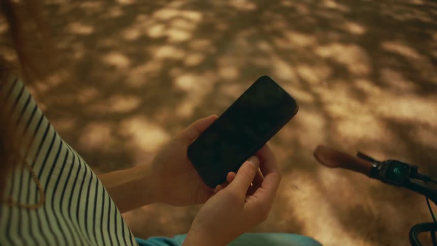 Over-the-shoulder close-up of a woman using her smartphone while sitting next to her bicycle in the park on a sunny day