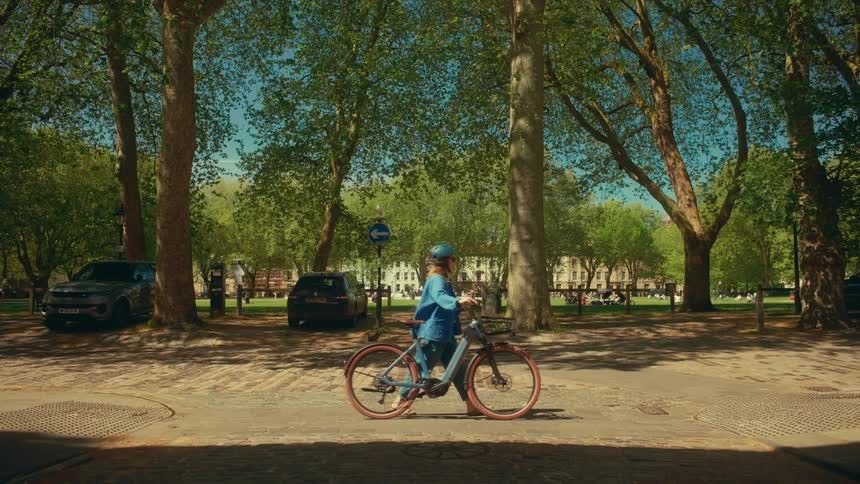 A woman walks her e-bike across a cobblestone street next to a sunlit urban park