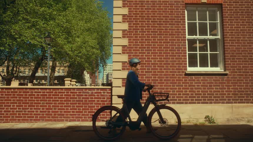 A woman wearing a helmet walks her e-bike along a sunny city sidewalk past a red brick building