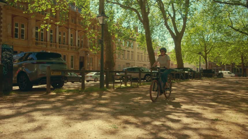 A woman wearing a helmet rides her bicycle on a dirt path through a sun-dappled city park