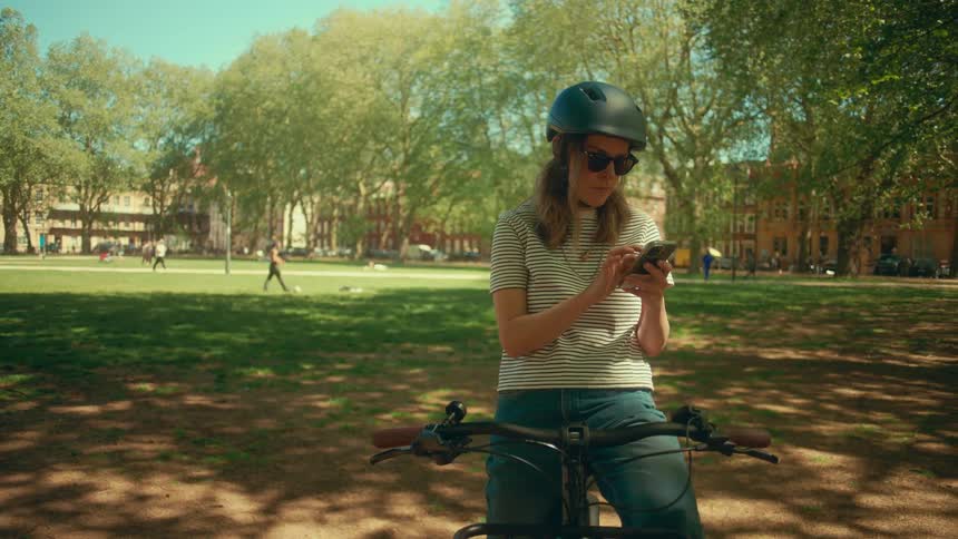 A woman wearing a bike helmet and sunglasses scrolls on her smartphone while standing with her bicycle in a sunny city park