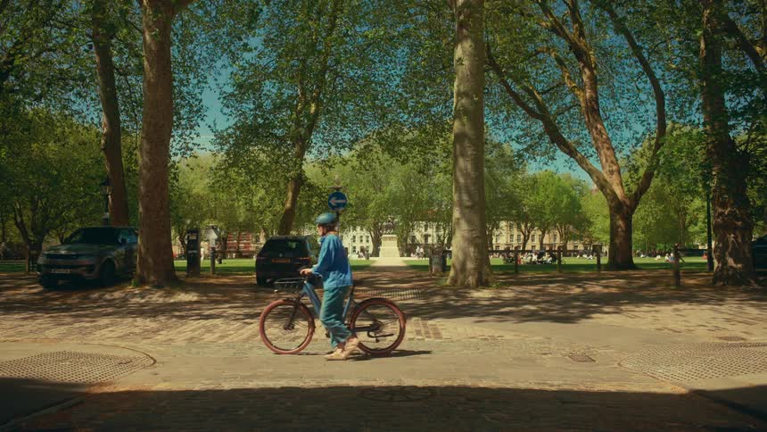 Slow motion shot of a woman walking her bicycle across a sun-dappled cobblestone street with a lush city park in the background