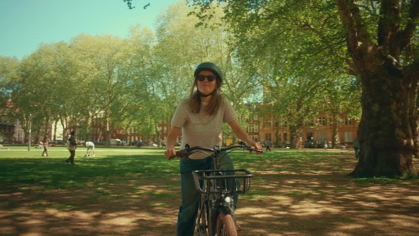 A woman wearing a helmet and sunglasses checks her phone before happily riding her electric bicycle through a sunny city park
