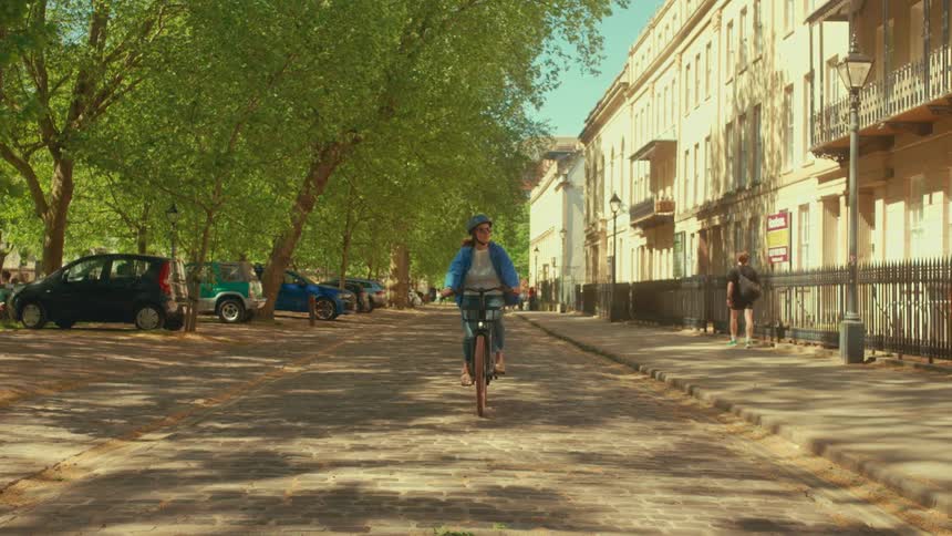 A woman wearing a helmet and sunglasses happily rides her bicycle towards the camera down a sunny, tree-lined cobblestone street