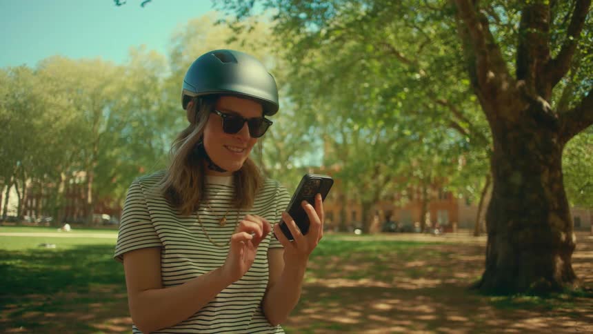 A smiling woman in a bicycle helmet and sunglasses scrolls on her smartphone in a sunlit city park