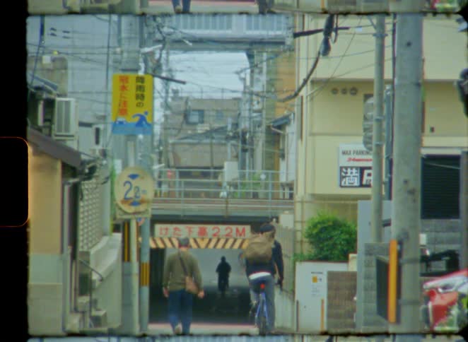 A vintage 8mm film captures people walking and cycling down a narrow street into an underpass in a Japanese city