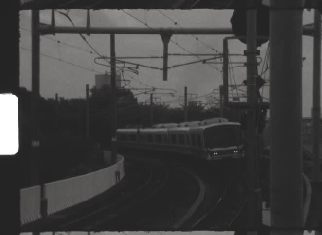 A vintage black and white film captures a modern passenger train approaching on a curving railroad track
