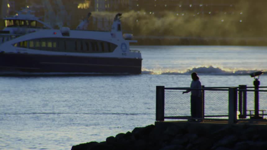 A silhouetted person stands on a pier watching an NYC ferry pass by on the water during a beautiful golden hour