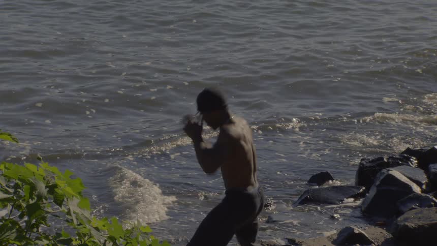A muscular, shirtless man in a beanie intensely shadowboxes at the water's edge on a rocky shore