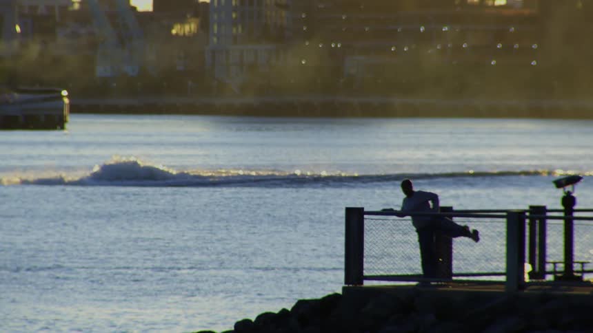 A silhouetted person stretches on a pier railing by the water during a hazy golden hour