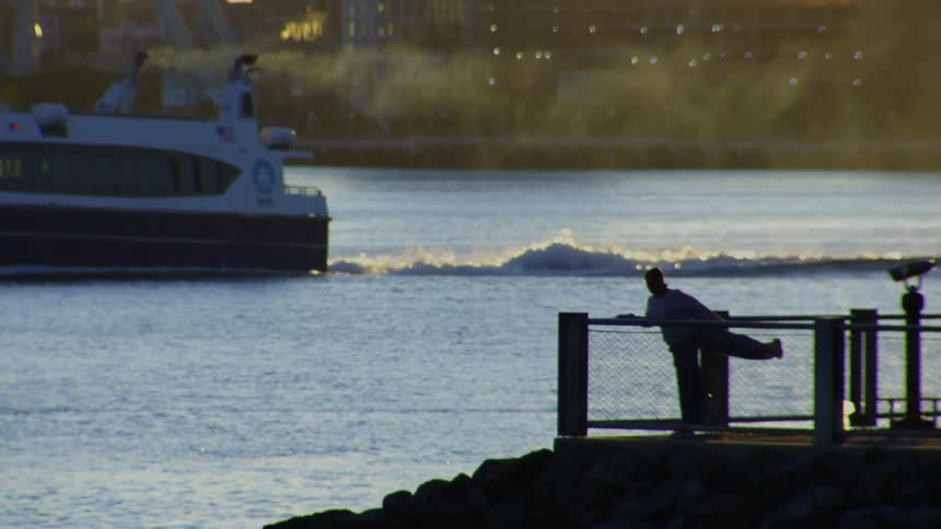 A silhouetted person stretches on a city pier at golden hour as a water taxi cruises by in the background