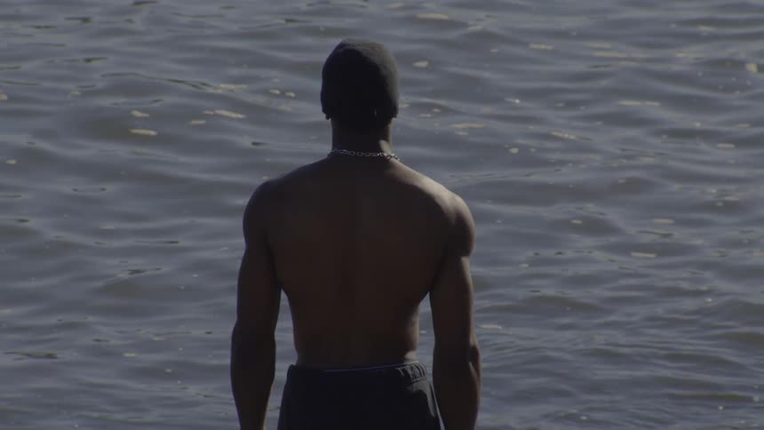 Rear view of a muscular, shirtless man in a beanie standing still and looking out at the water
