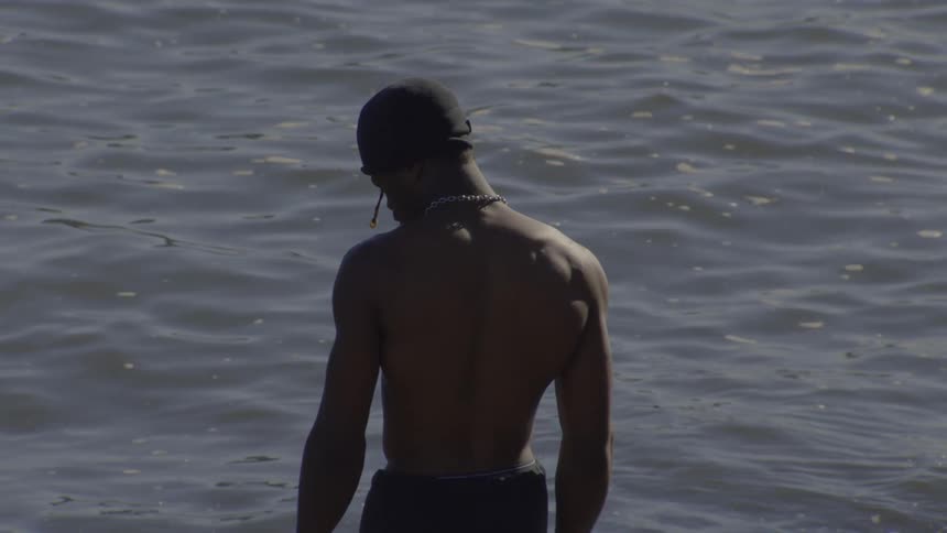 A muscular man in a beanie slowly turns around with a smile while standing in front of a body of water
