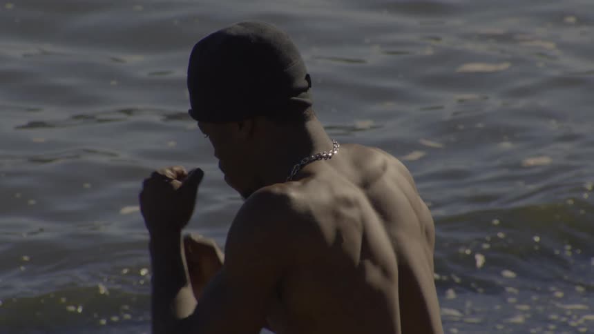 A muscular shirtless man with his back to the camera practices slow-motion boxing punches while standing in the water