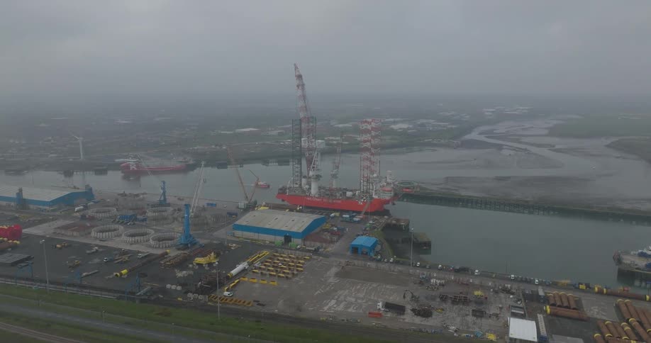 A wide static aerial view of a huge red offshore construction vessel docked at an industrial harbor on a misty, overcast day