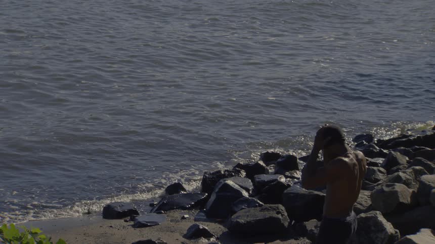 A shirtless, muscular man takes a moment to reflect by the water's edge on a rocky shore