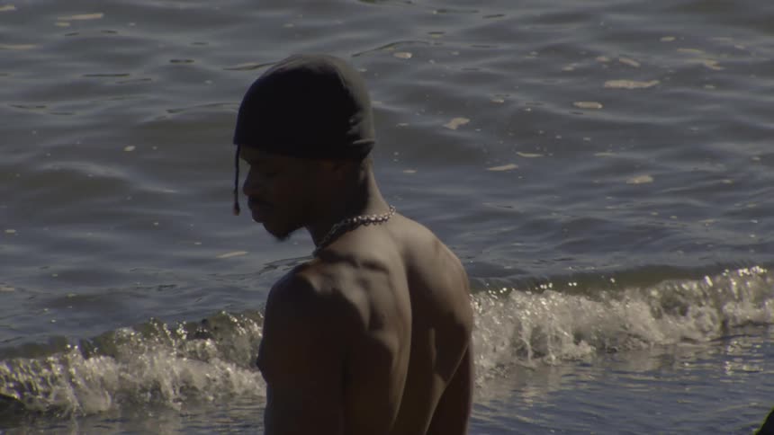 A muscular, shirtless man wearing a beanie stands in the water and turns to look back at the camera in slow motion as waves roll in