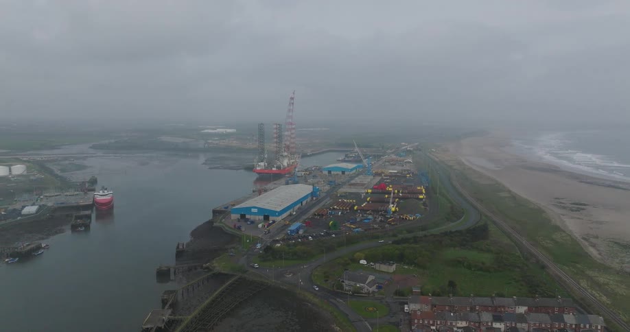 A wide aerial view of a large coastal shipyard with docked vessels and industrial equipment on a misty, overcast day