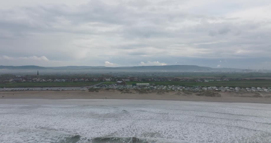 An aerial view from the sea pushes toward a sandy beach and coastal town on a cloudy, misty day