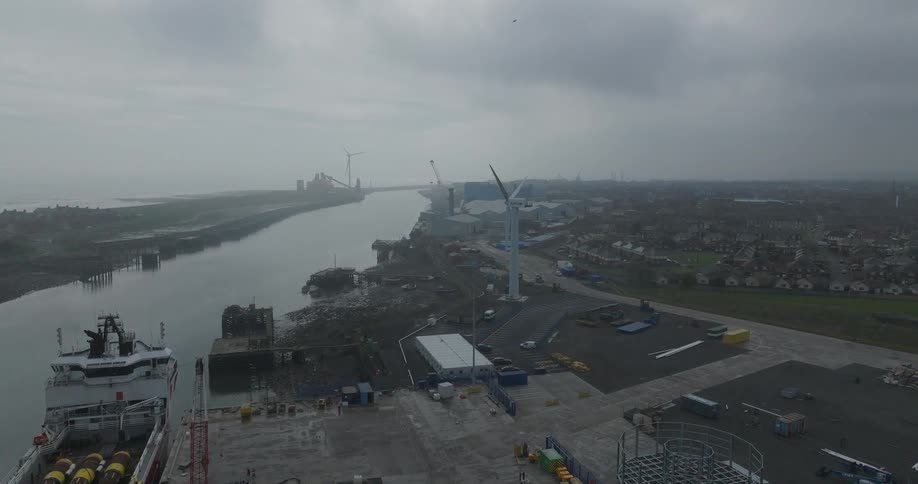 An aerial view of a large industrial port showing cargo ships docked and wind turbines under construction on a foggy, overcast day