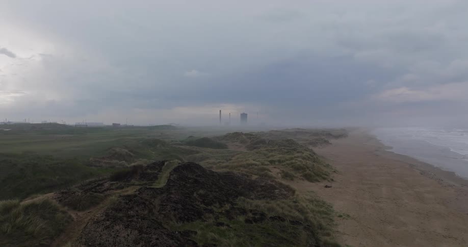 Slow-motion aerial footage shows a seagull flying over a foggy, desolate beach with a looming industrial plant in the background
