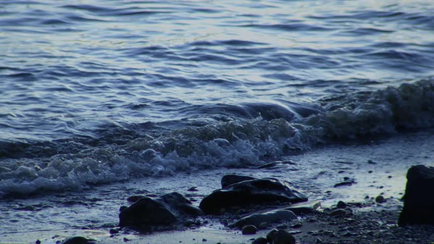 Gentle blue waves wash over a rocky shoreline in slow motion